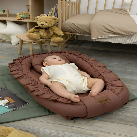 Baby lying on a brown ruffled baby seat in a room with a teddy bear and books.